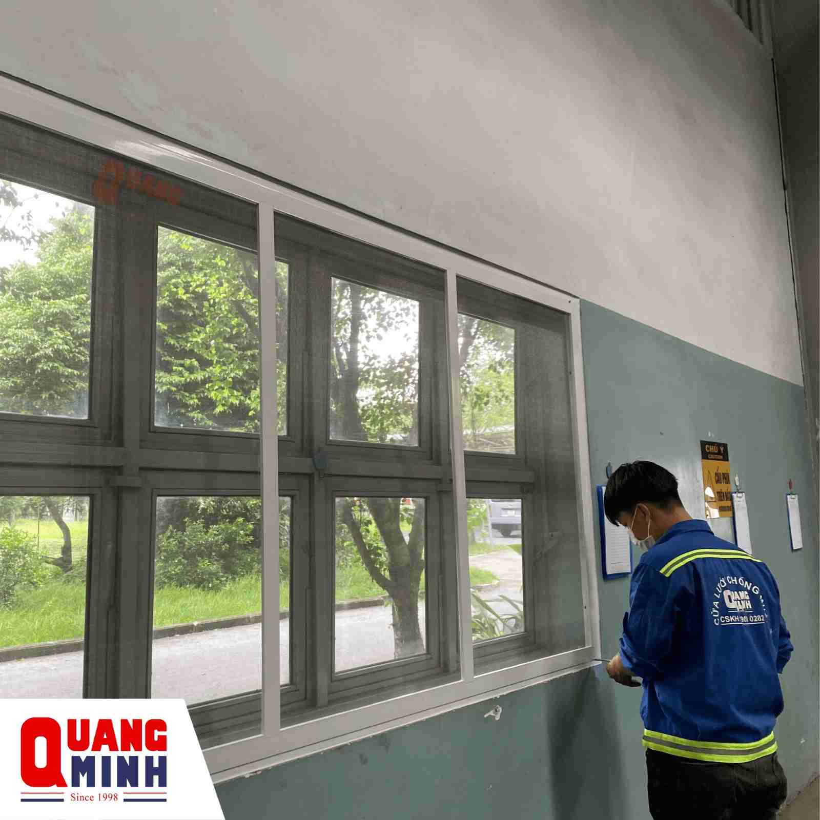 Vách lưới cố định lắp tại cửa sổ, có khung trắng. Nhân viên Quang Minh đang kiểm tra vách lưới