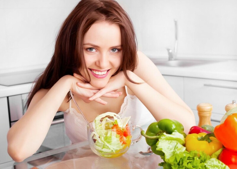 Người phụ nữ đang ngồi tươi cười trước bàn ăn đầy các loại rau xanh